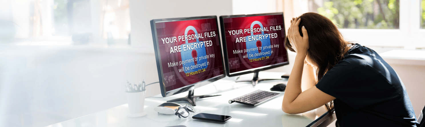 Frustrated woman holding her head in front of dual monitors displaying a ransomware message stating 'Your personal files are encrypted' with a countdown timer and payment demand.