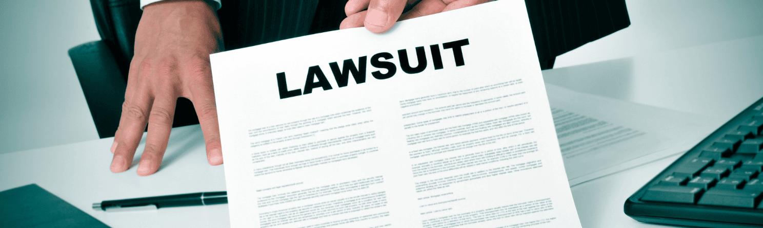 Close-up of a person in a suit holding a document titled 'LAWSUIT' over a desk with a keyboard and pen.