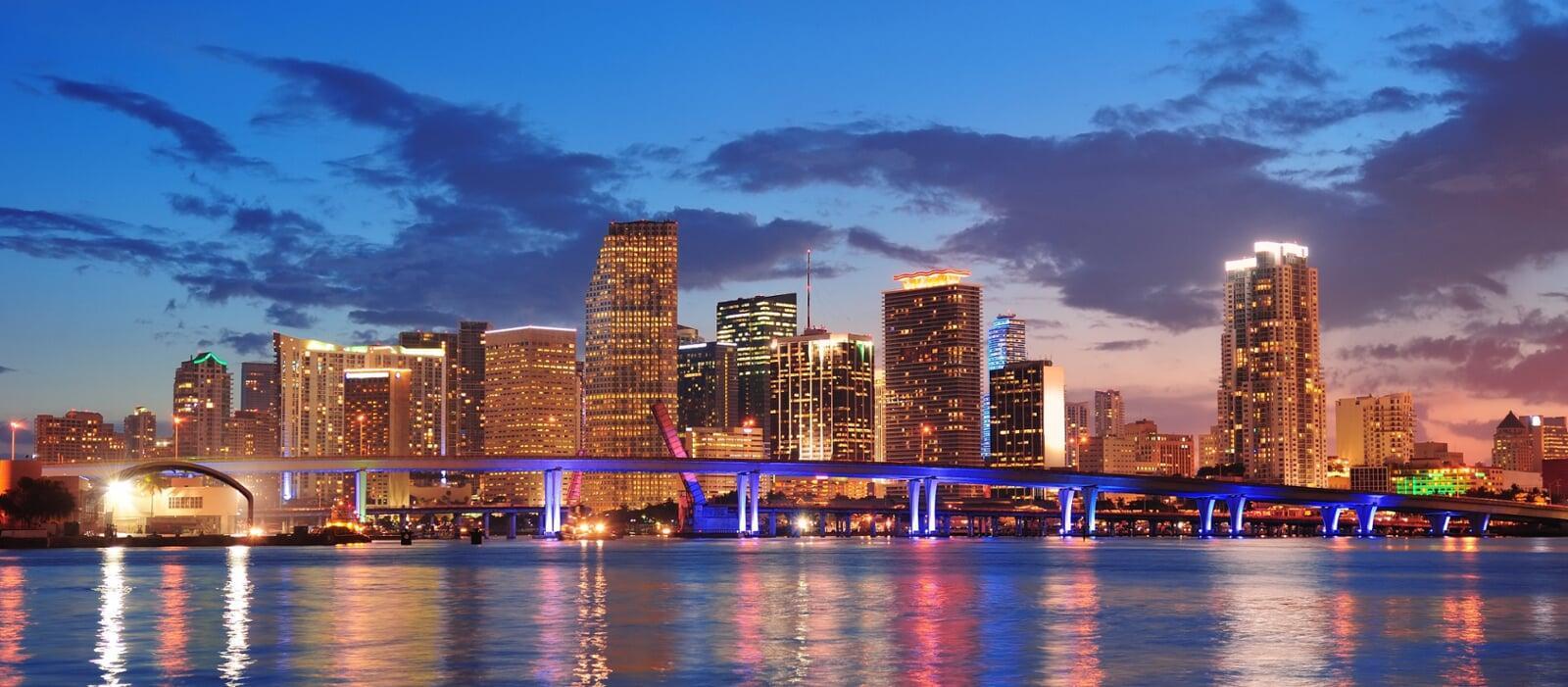 Miami skyline at dusk with illuminated skyscrapers, a scenic bridge, and city lights reflecting on the water