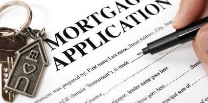 Close-up of a hand signing a mortgage application form with a house-shaped keychain beside it.